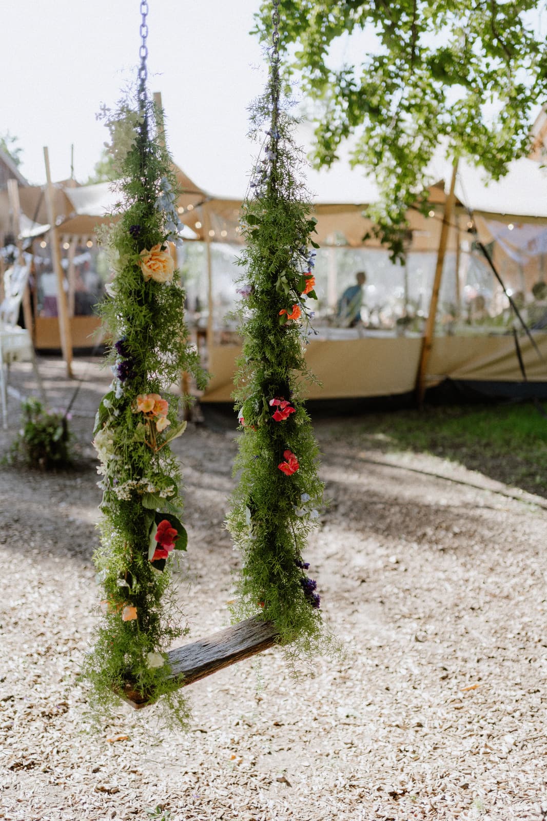 Mit Wildblumen und Grün geschmückte Holzschaukel vor dem Stretchzelt der Hochzeitsfeier